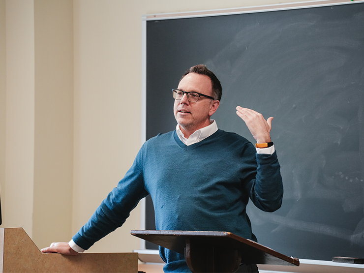 An instructors stands at a podium and speaks to a classroom.